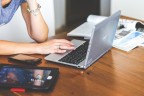 Une personne consulte son ordinateur, entourée d'un téléphone, d'une tablette et de journaux
